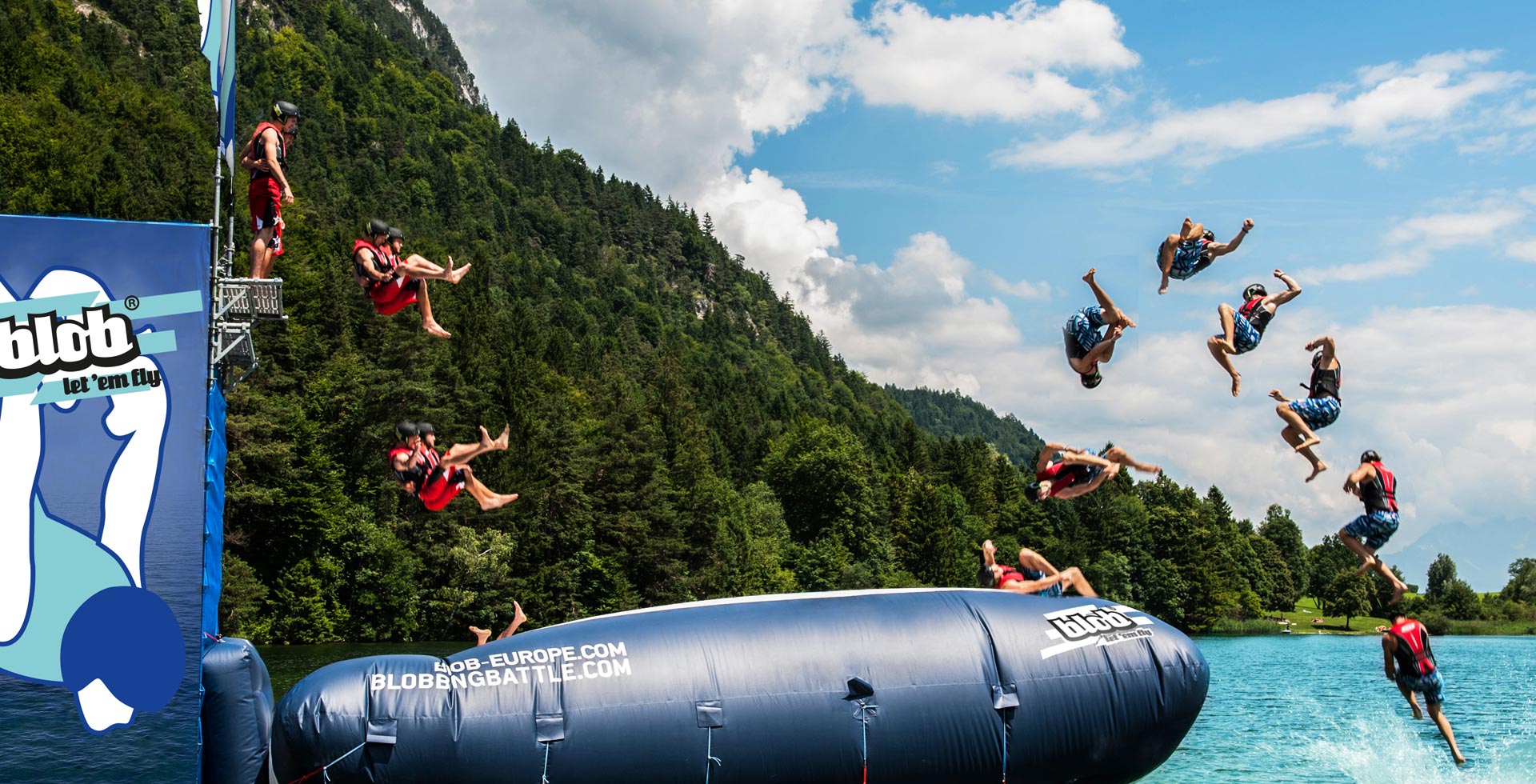 Menschen in Schwimmwesten werden von einem großen aufblasbaren Blob in einen See geworfen, mit bewaldeten Hügeln und Wolken im Hintergrund. Mehrere Action-Aufnahmen zeigen Saltos und Sprünge in der Luft.