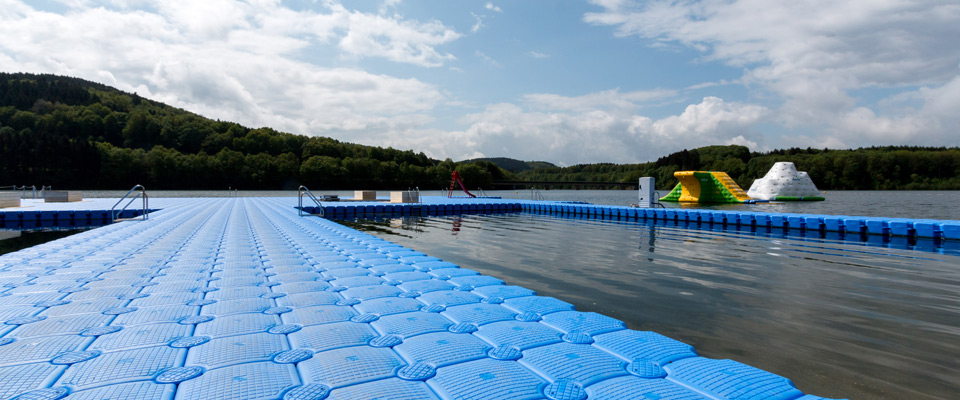 Ein großer blauer Schwimmsteg führt in einen See mit aufblasbaren Spielgeräten und Rutschen im Wasser, umgeben von grünen Bäumen und Hügeln unter einem teilweise bewölkten Himmel.